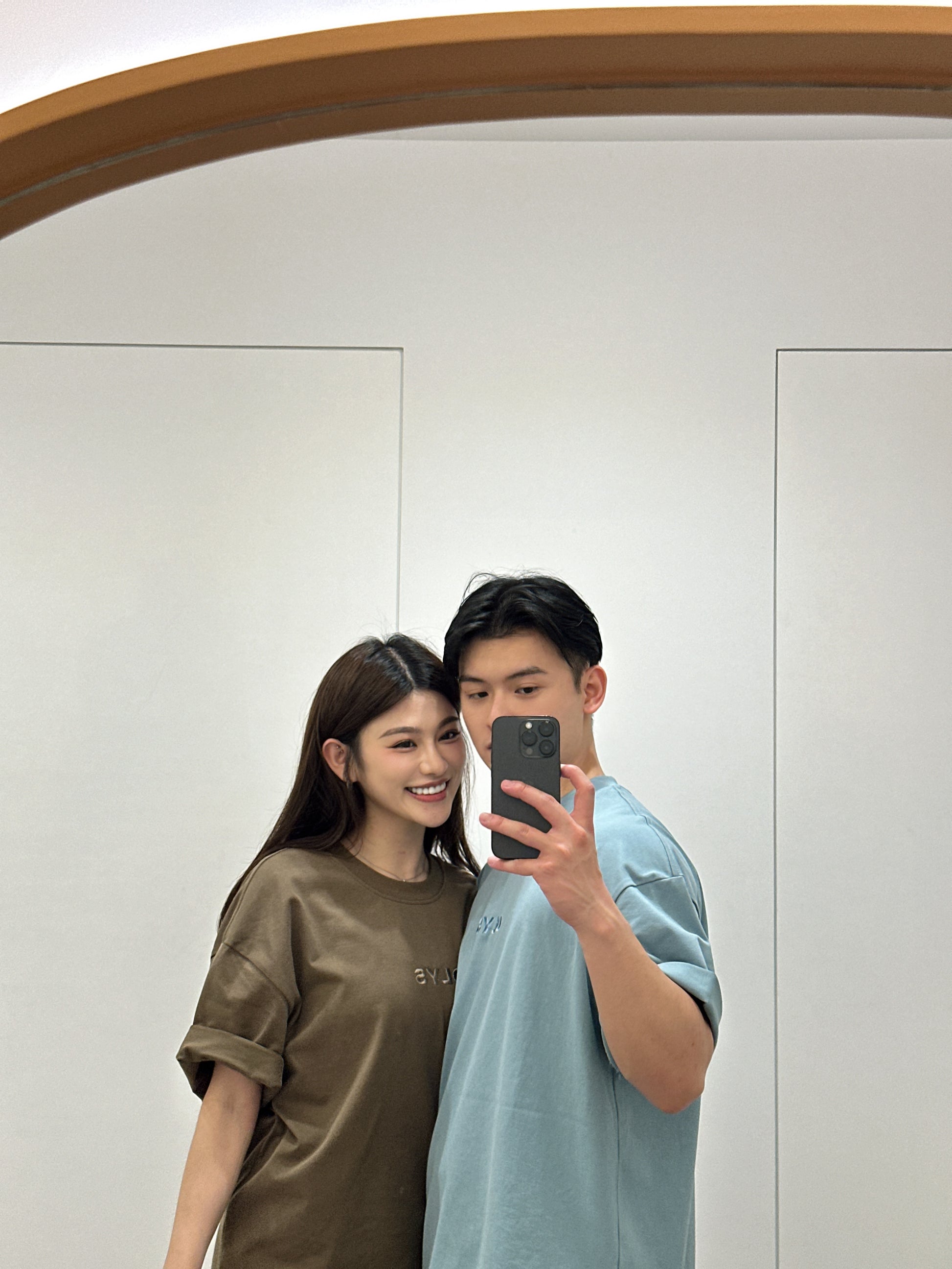 情侶穿搭自拍,女生穿著棕色 OLYS 刺繡 Logo 寬鬆 T 恤,男生穿著淺藍色 OLYS 刺繡 Logo 寬鬆 T 恤,簡約休閒風格,鏡子自拍