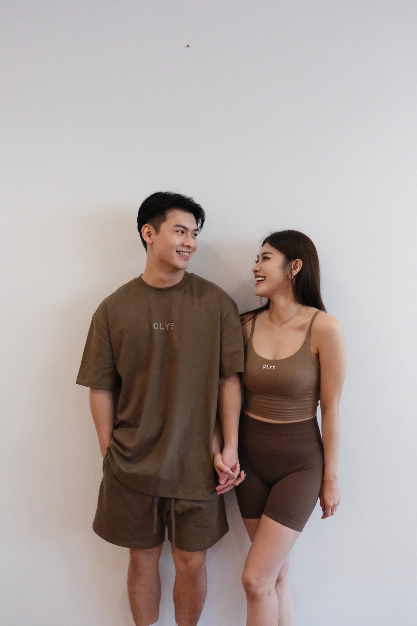 情侶穿搭,男生穿著棕色 OLYS 刺繡 Logo 寬鬆 T 恤與短褲,女生穿著棕色 OLYS 美背運動背心搭配高腰翹臀短褲,簡約舒適的情侶風格